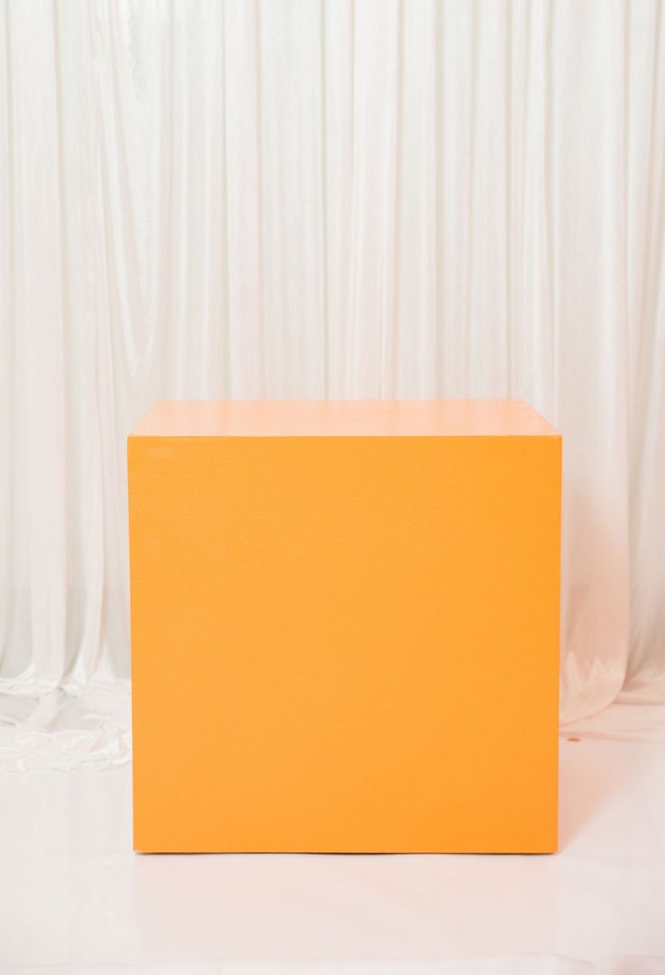 A yellow cube is sitting in front of white curtains.